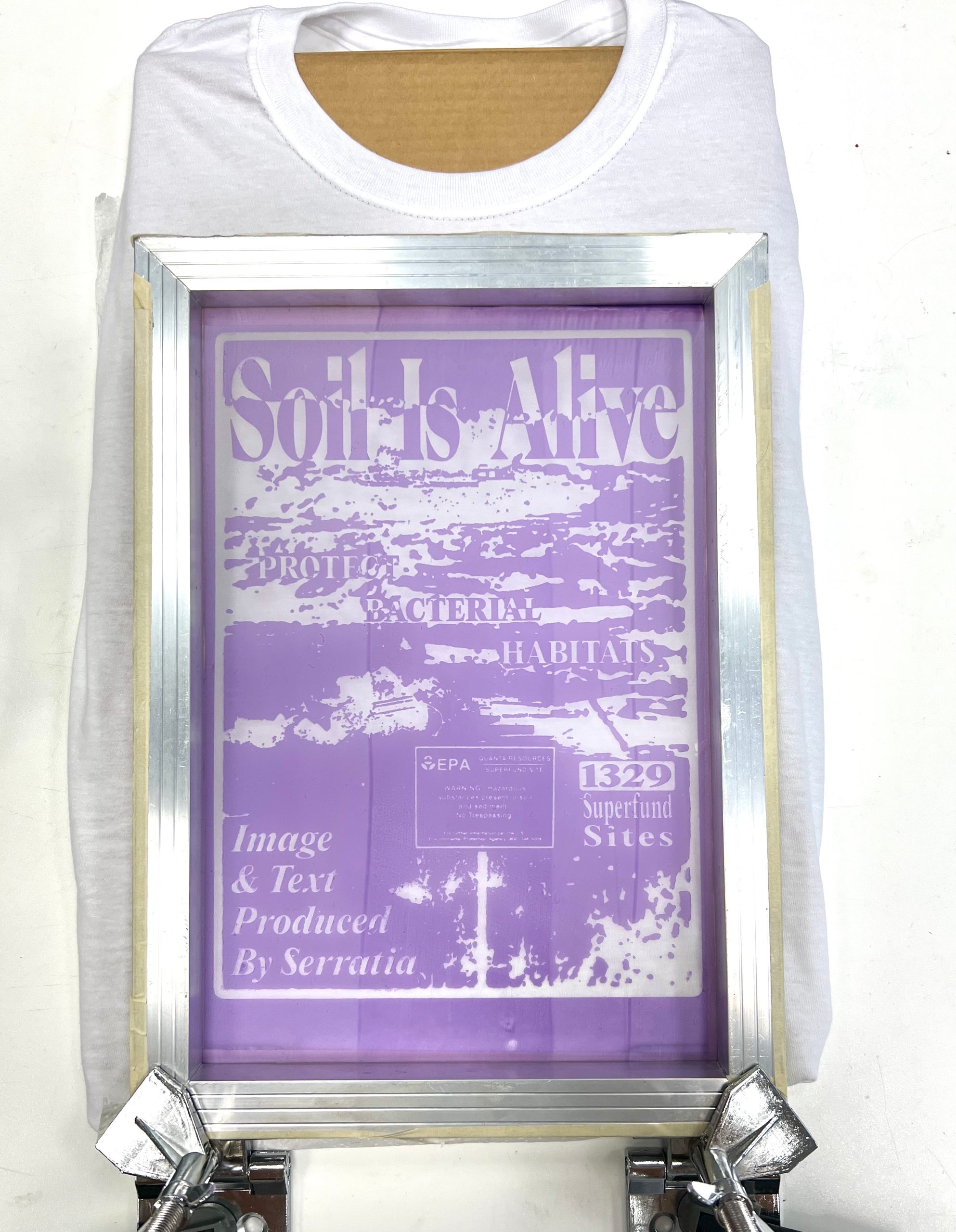 A screen-printing frame clamped over a white t-shirt, the “Soil Is Alive” design showing through the mesh in pale purple.