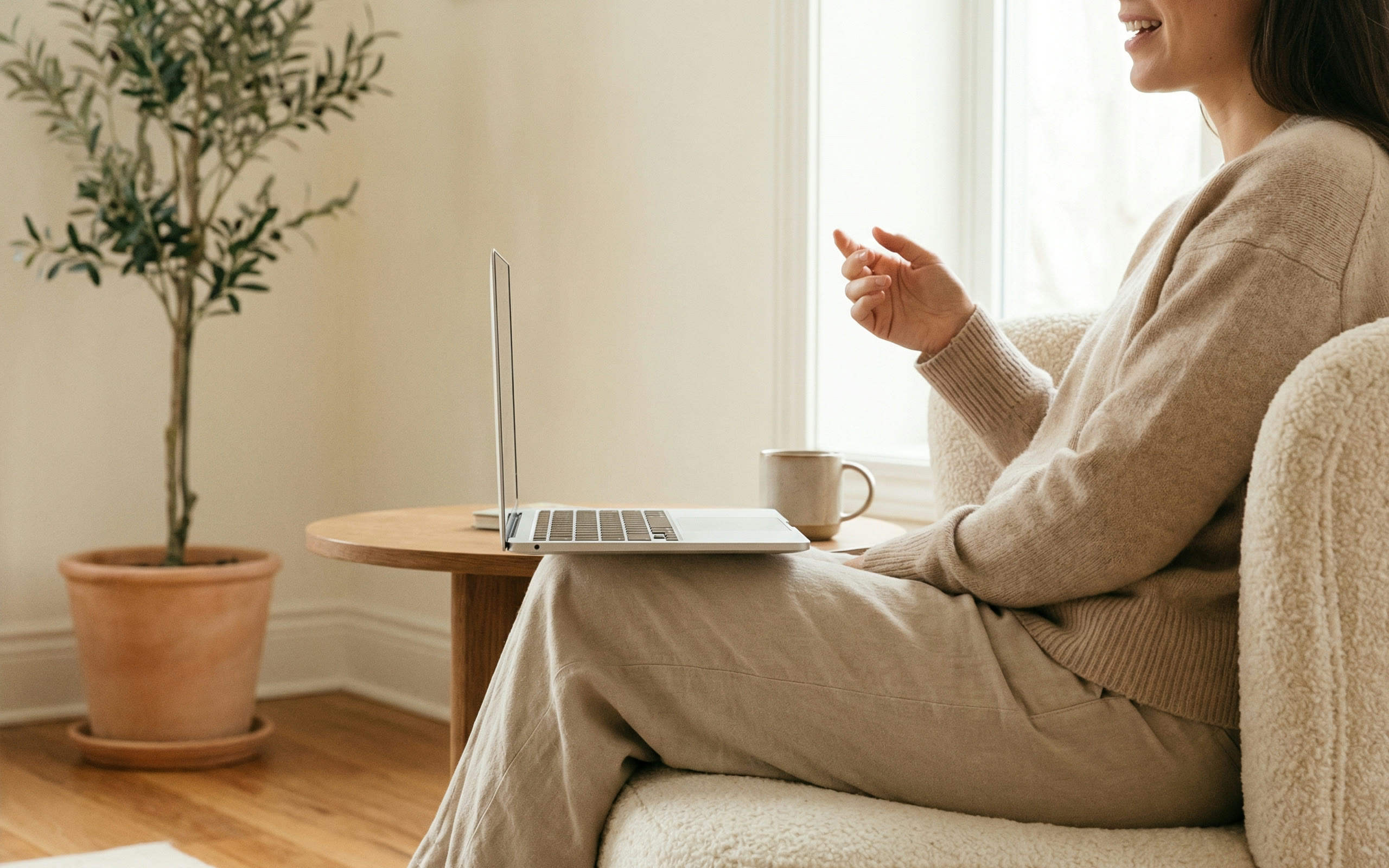 A woman on a cream sofa talking to a laptop on a side table, an olive tree in a pot beside the window.