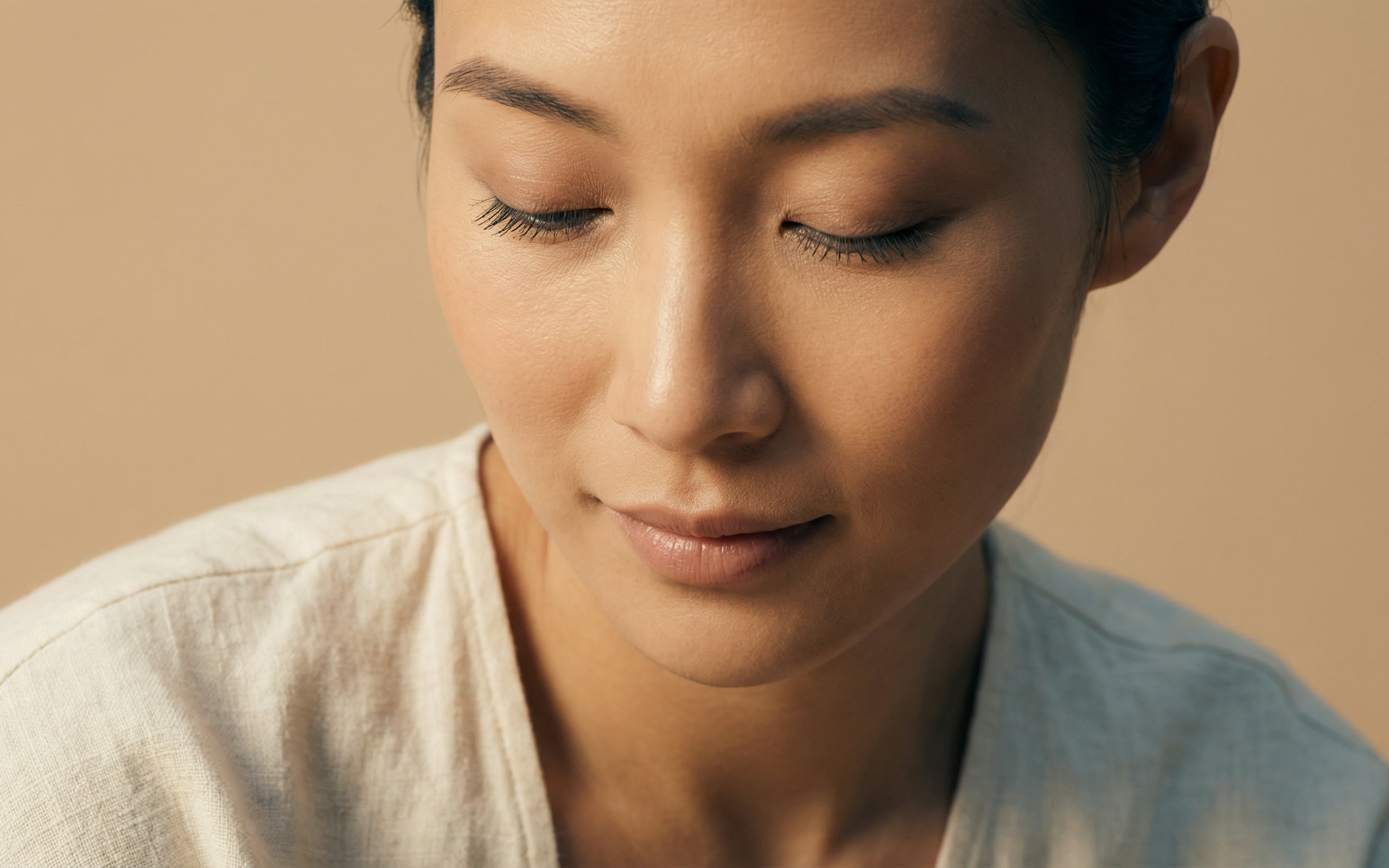 A close portrait of a woman with her eyes lowered, in a linen top against a warm sand wall.