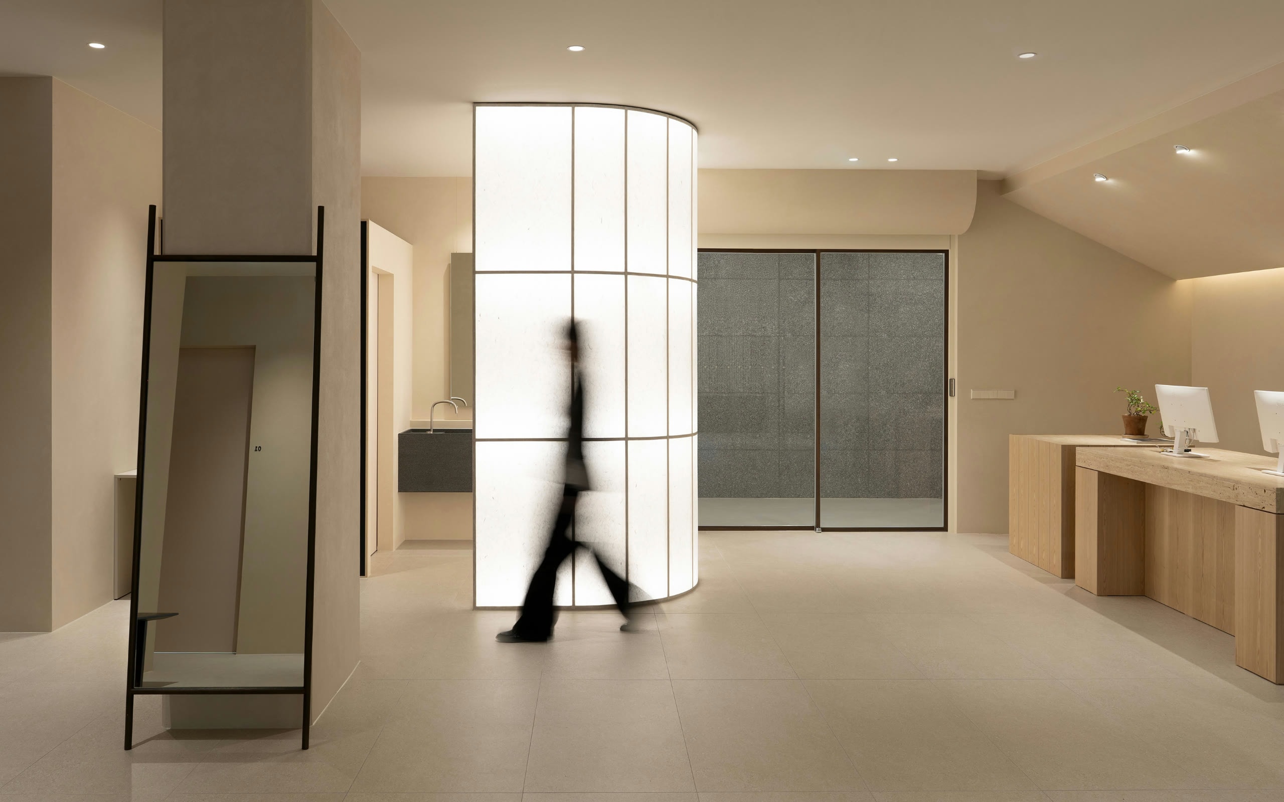 A pale clinic lobby with a lit glass column and a blurred figure walking past it.