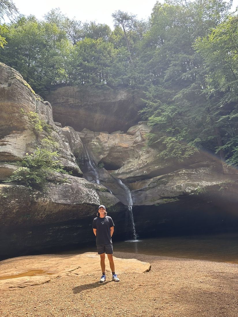Gabe standing small at the foot of a huge sandstone cave with a thin waterfall.