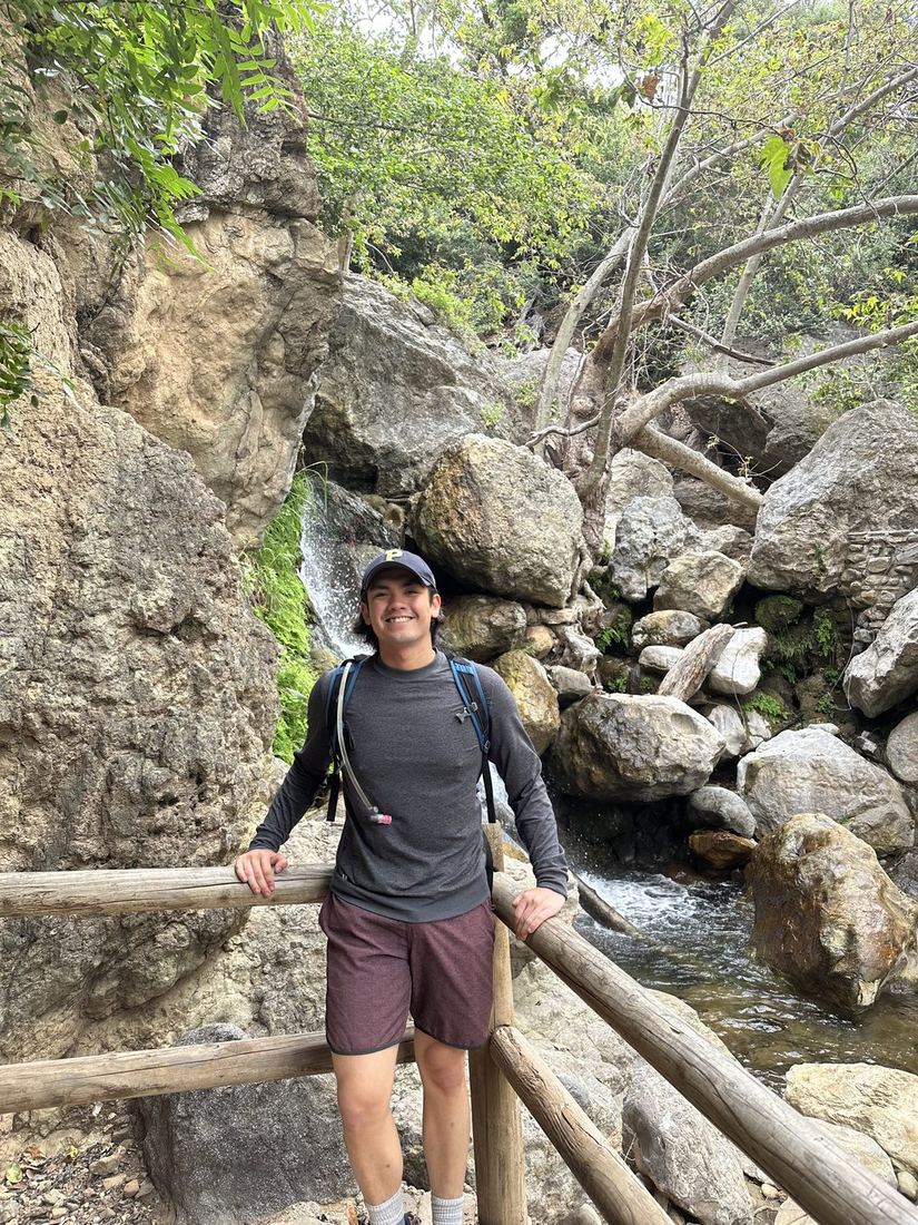 Gabe leaning on a wooden rail on a rocky, shaded trail.